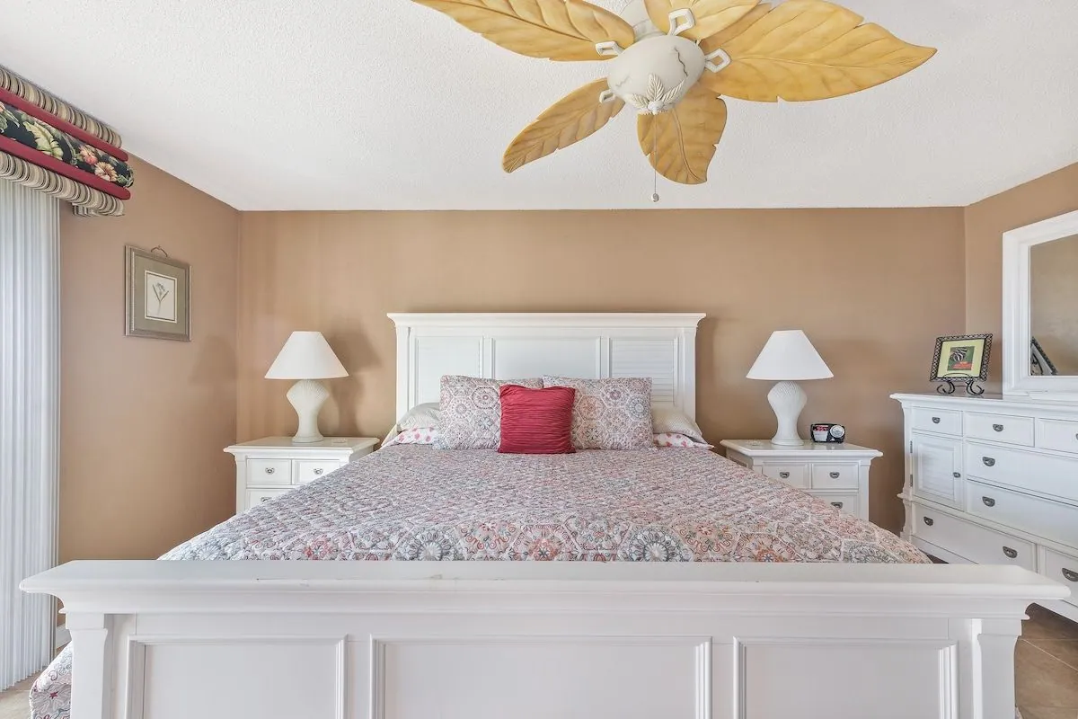 King bedroom with white coastal furniture, paired nightstands and lamps, and a palm-leaf ceiling fan