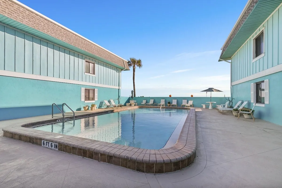 Heated outdoor pool in the center of the villa building, framed by teal walls, sun loungers, a patio umbrella and a palm tree